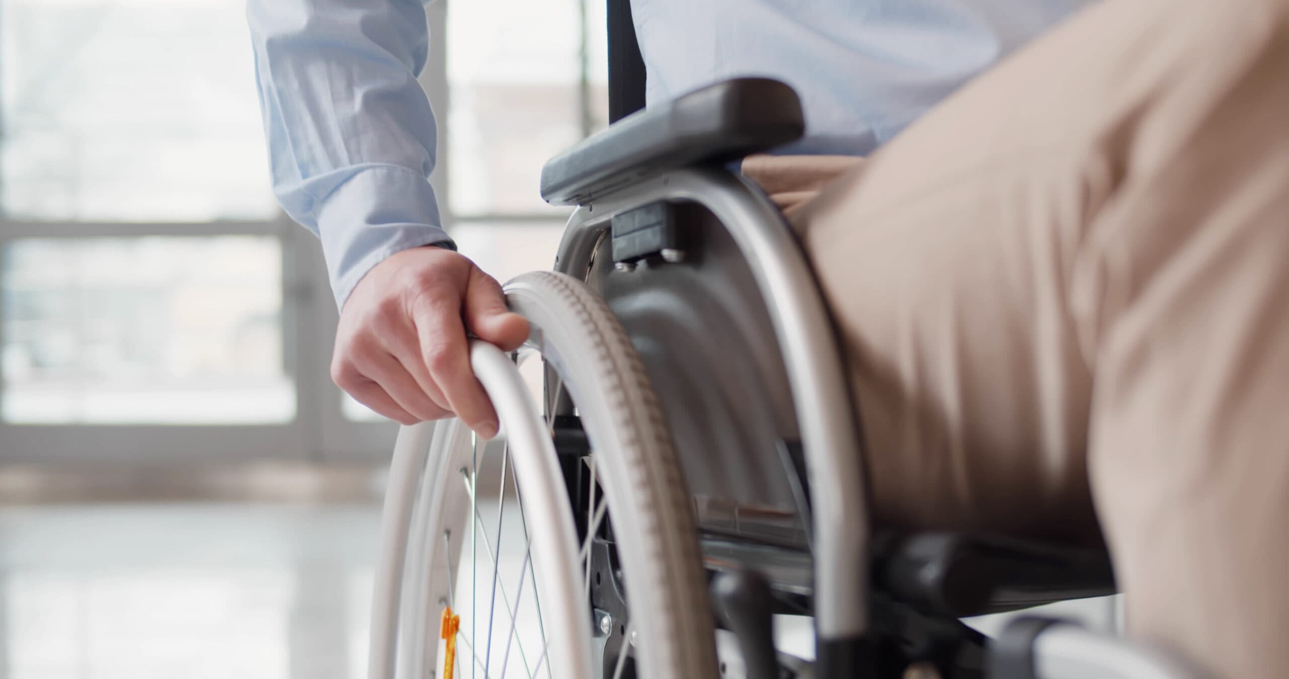 Close up of disabled man riding in wheelchair. Cropped shot of handicapped male holding hand on wheel of wheelchair indoors. Office worker in wheelchair in business center